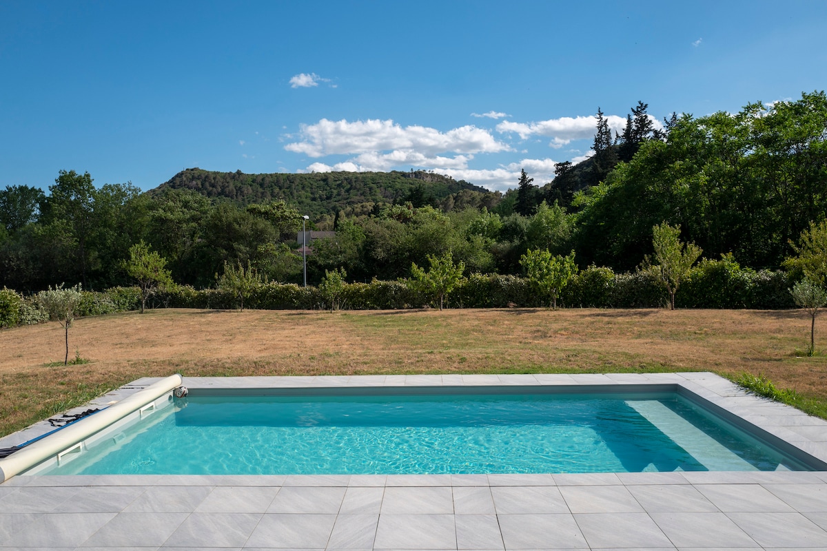 A tranquil view of a swimming pool is captured, showcasing clear water reflecting the sky. Lush greenery and rolling hills surround the pool area, providing a serene backdrop. The poolside is paved with light-colored tiles, complementing the natural environment.