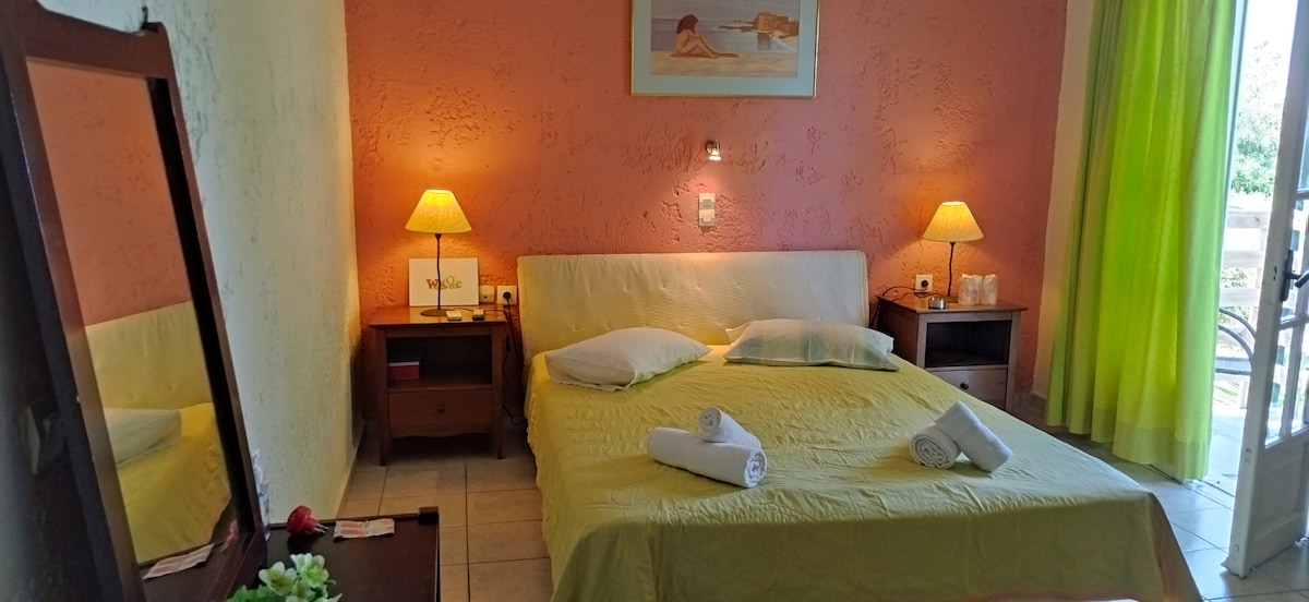 A bright sleeping area features a spacious bed with light yellow bedding, complemented by two pillows. Soft lighting is provided by two bedside lamps. A mirror reflects the room, while large windows with light green curtains allow natural light to fill the space.