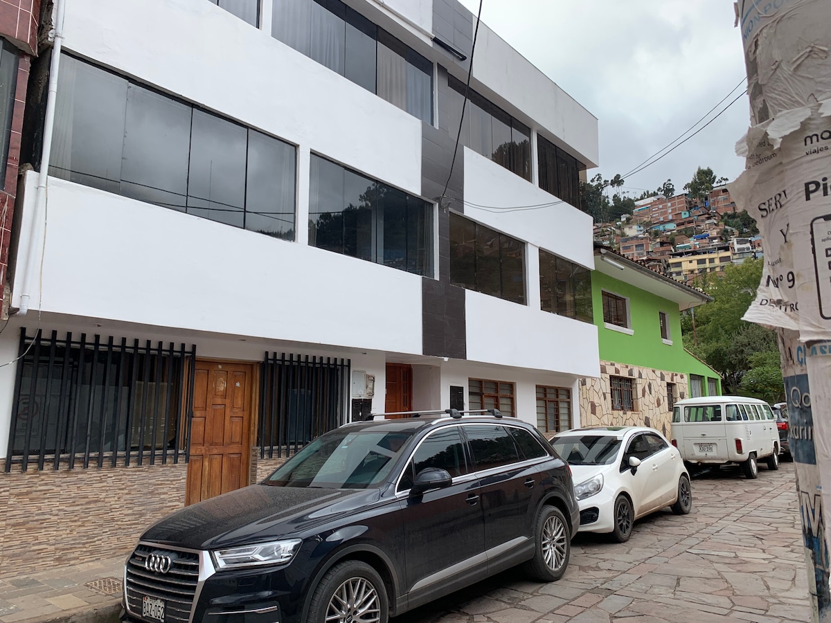 A modern building is featured with a mixture of white and dark exterior panels. Large glass windows reflect the street below, showcasing a vibrant neighborhood. Several vehicles are parked along the cobblestone road in front of the structure, contributing to an urban atmosphere.