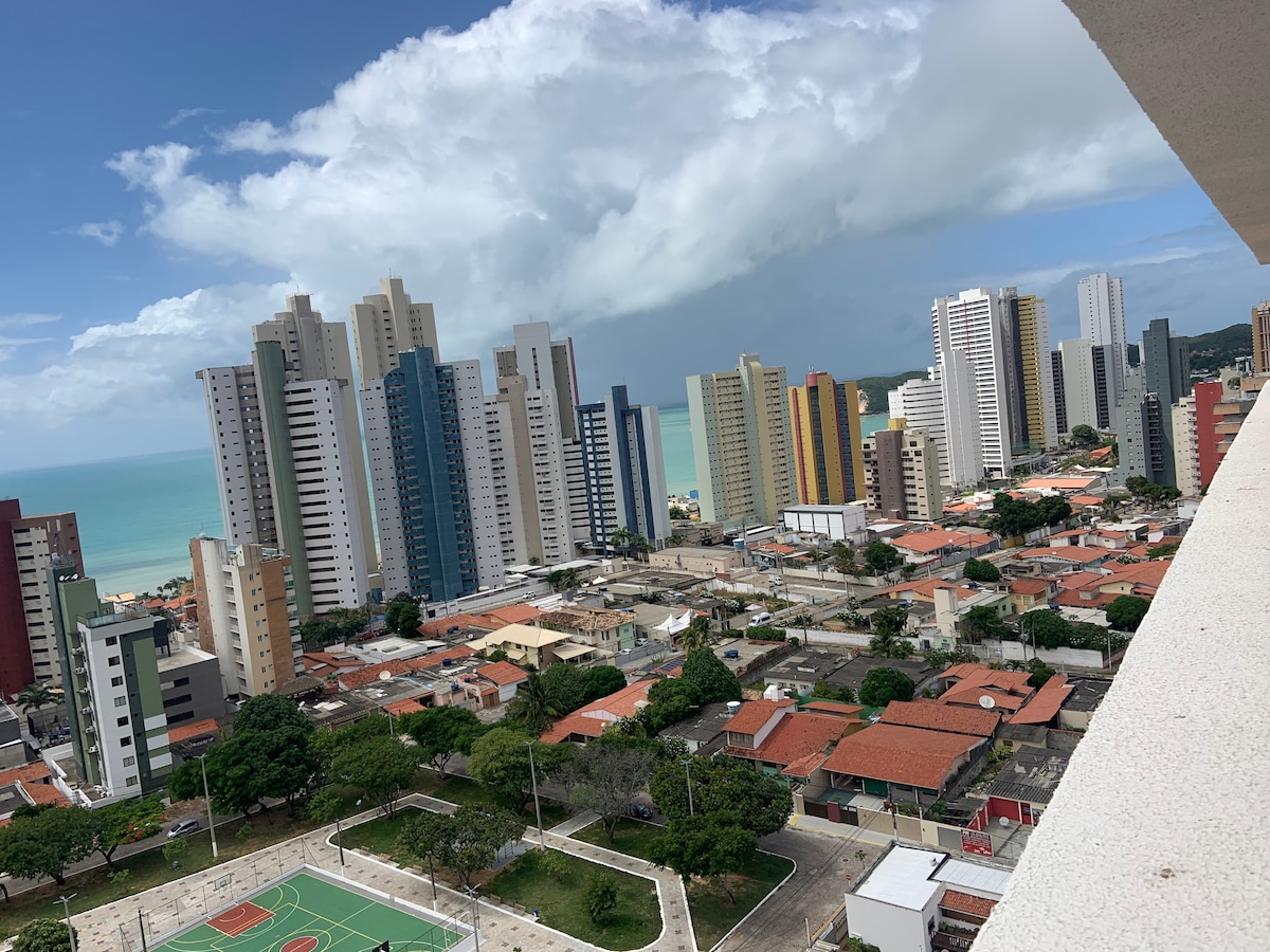 A panoramic view captures a vibrant urban landscape with high-rise buildings, showcasing a blend of modern architecture and residential areas. The coastline is visible in the distance against a backdrop of blue sky and scattered clouds, while a park area with a basketball court sits below.