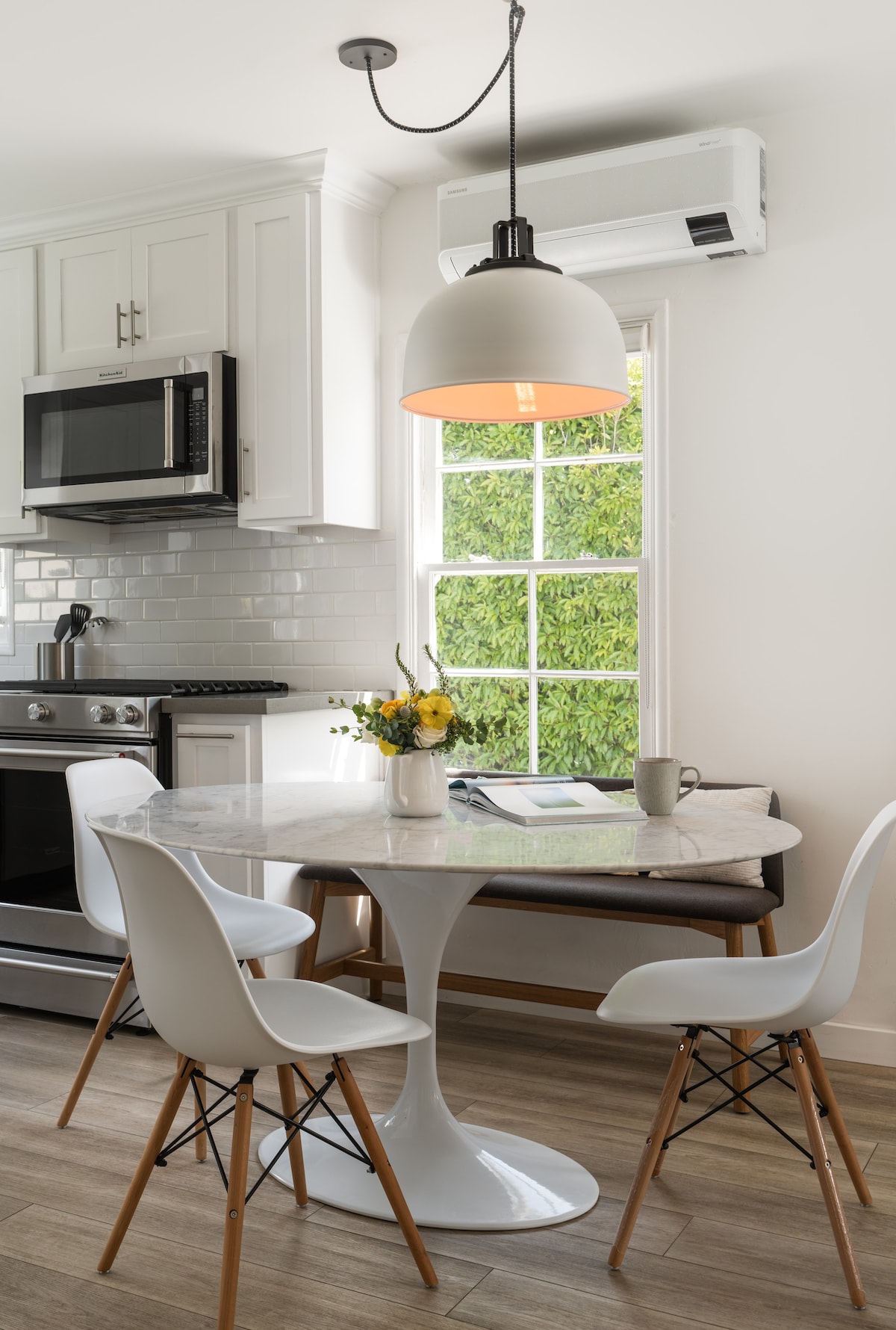 A modern kitchen area features a round marble dining table with four white chairs. Natural light filters through the window, highlighting a vase of flowers on the table. A sleek microwave and stainless steel stove are visible, complemented by a subtle pendant light overhead.