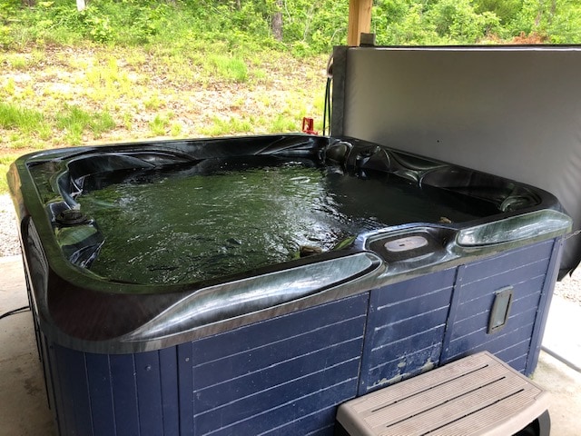 A private hot tub is situated under a covered area, featuring a spacious design with dark edges and clear water. Surrounding greenery is visible in the background, providing a natural view.