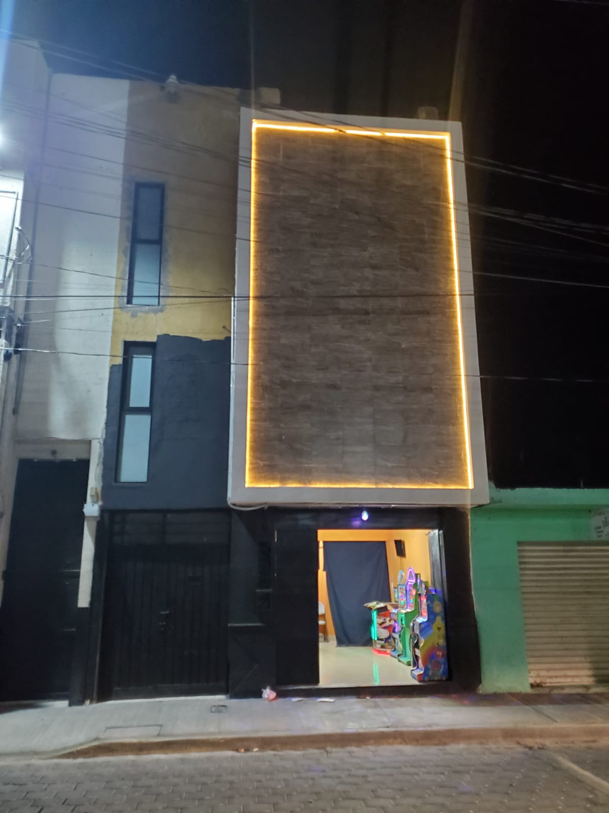 A modern building facade is illuminated by a subtle light strip along the vertical edges. The entrance is framed by a minimalist design, revealing glimpses of colorful decor inside through the open door. A dark exterior contrasts with the bright surroundings.
