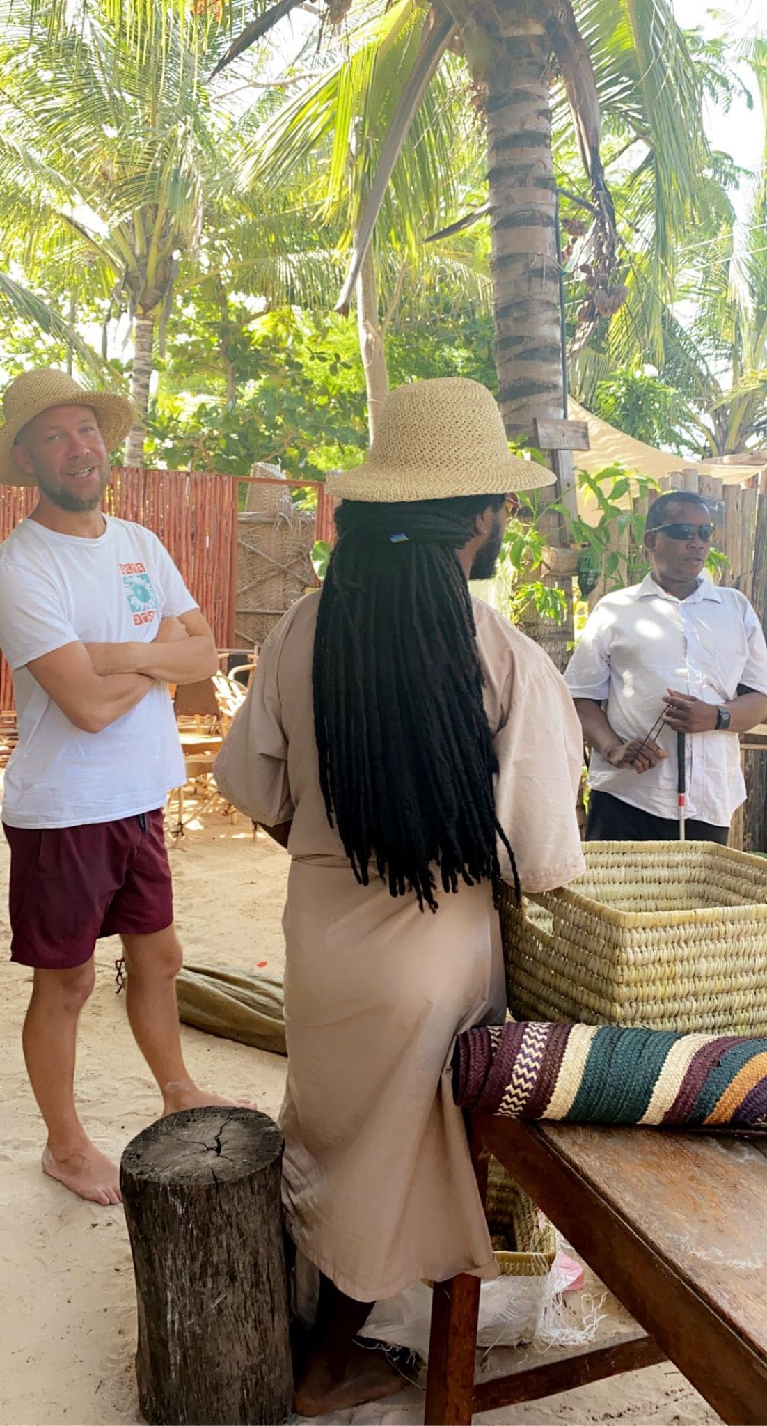 Two individuals stand near a picnic-style table surrounded by palm trees. One person, viewed from behind, wears a long, beige garment and a straw hat. Conversations appear to be taking place in a relaxed outdoor setting.