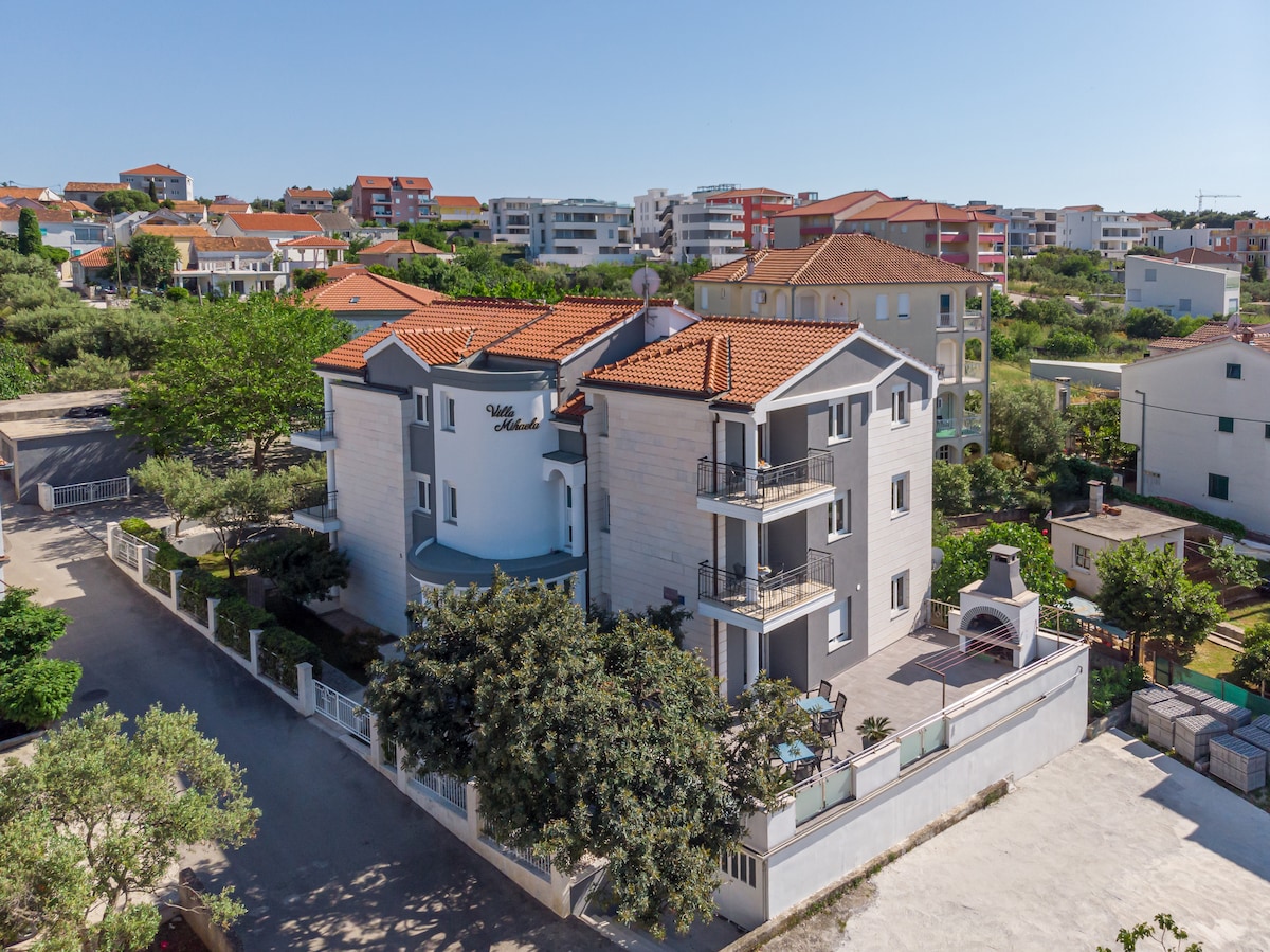 The Villa Mihaela is showcased from an elevated perspective, highlighting its modern architecture and terracotta roof. Lush greenery surrounds the property, with a barbecue area visible in the outdoor space. Nearby buildings and a clear blue sky create a serene backdrop.