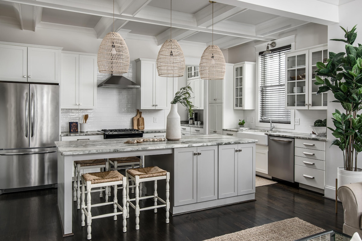 A modern kitchen features white cabinetry with a marble countertop and stainless steel appliances. Three woven pendant lights hang above a central island with two high stools. A large window allows natural light to illuminate the space, complemented by greenery from a nearby potted plant.