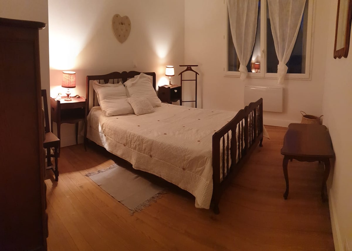 A well-lit bedroom features a spacious bed with light bedding and decorative pillows. Nightstands with lamps are positioned on either side, casting a soft glow. A vintage dresser stands nearby, and a warm wooden floor complements the neutral decor. Windows adorned with sheer curtains offer gentle illumination.