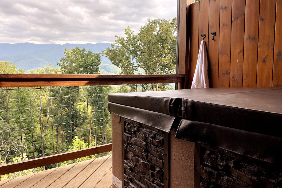 A hot tub is situated on a wooden deck, surrounded by a rustic wood railing. Expansive mountain views are visible in the background, framed by lush green trees under a partly cloudy sky.