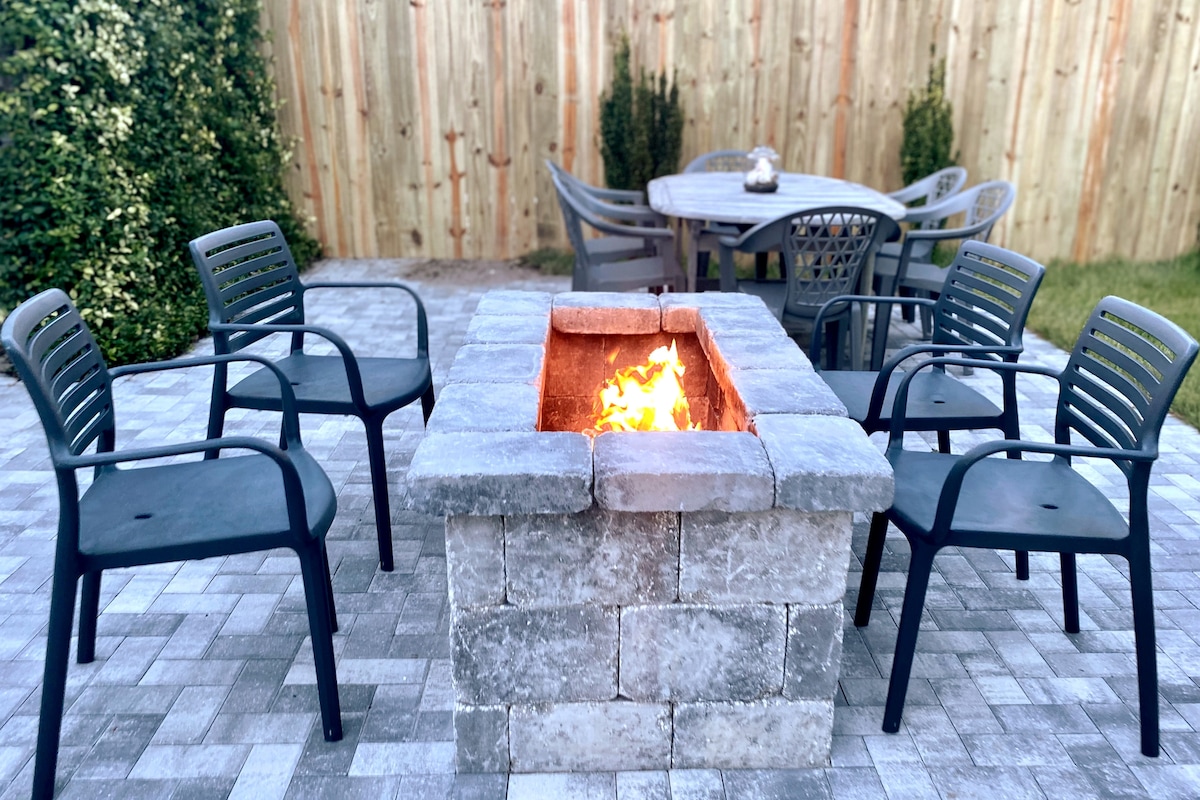 An outdoor patio area features a stone fire pit surrounded by six black chairs. A wooden table stands in the background, providing additional seating options. The setting is enclosed by a wooden fence and greenery, enhancing the outdoor ambiance.