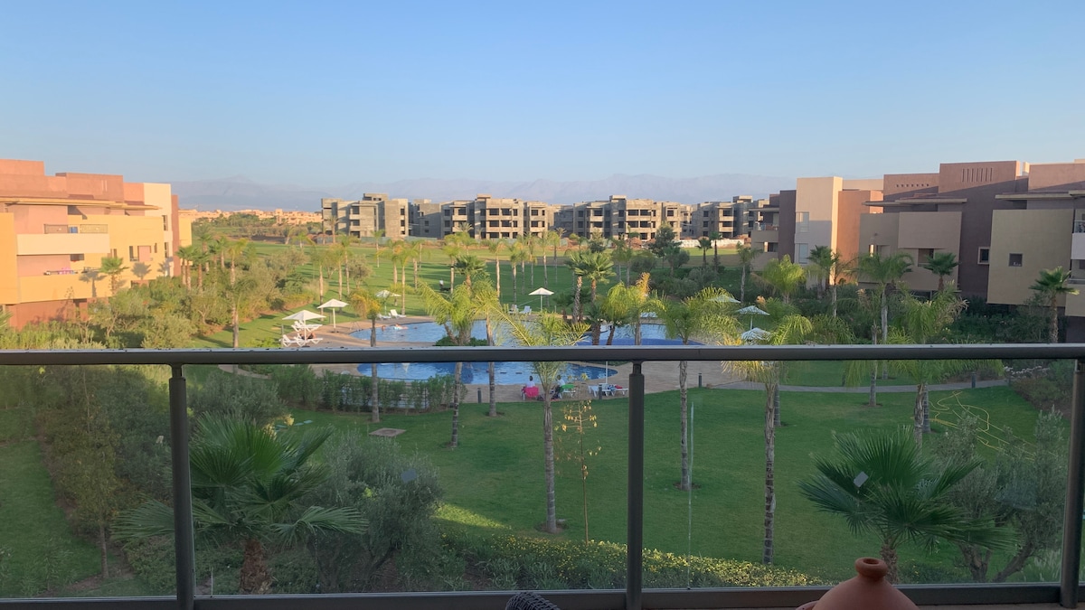 A spacious terrace overlooks a lush green area featuring palm trees and a pool. Surrounding buildings are visible in the background, showcasing a blend of modern architecture. The serene landscape extends towards distant mountains, enhancing the tranquil setting.