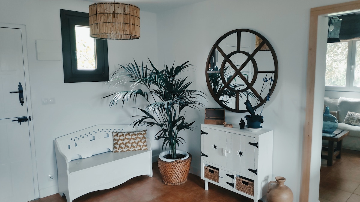 A welcoming entry area features a white bench adorned with patterned cushions alongside a potted palm plant. A round mirror is positioned above a white cabinet with star motifs and storage baskets, contributing to the light and airy feel of the space.