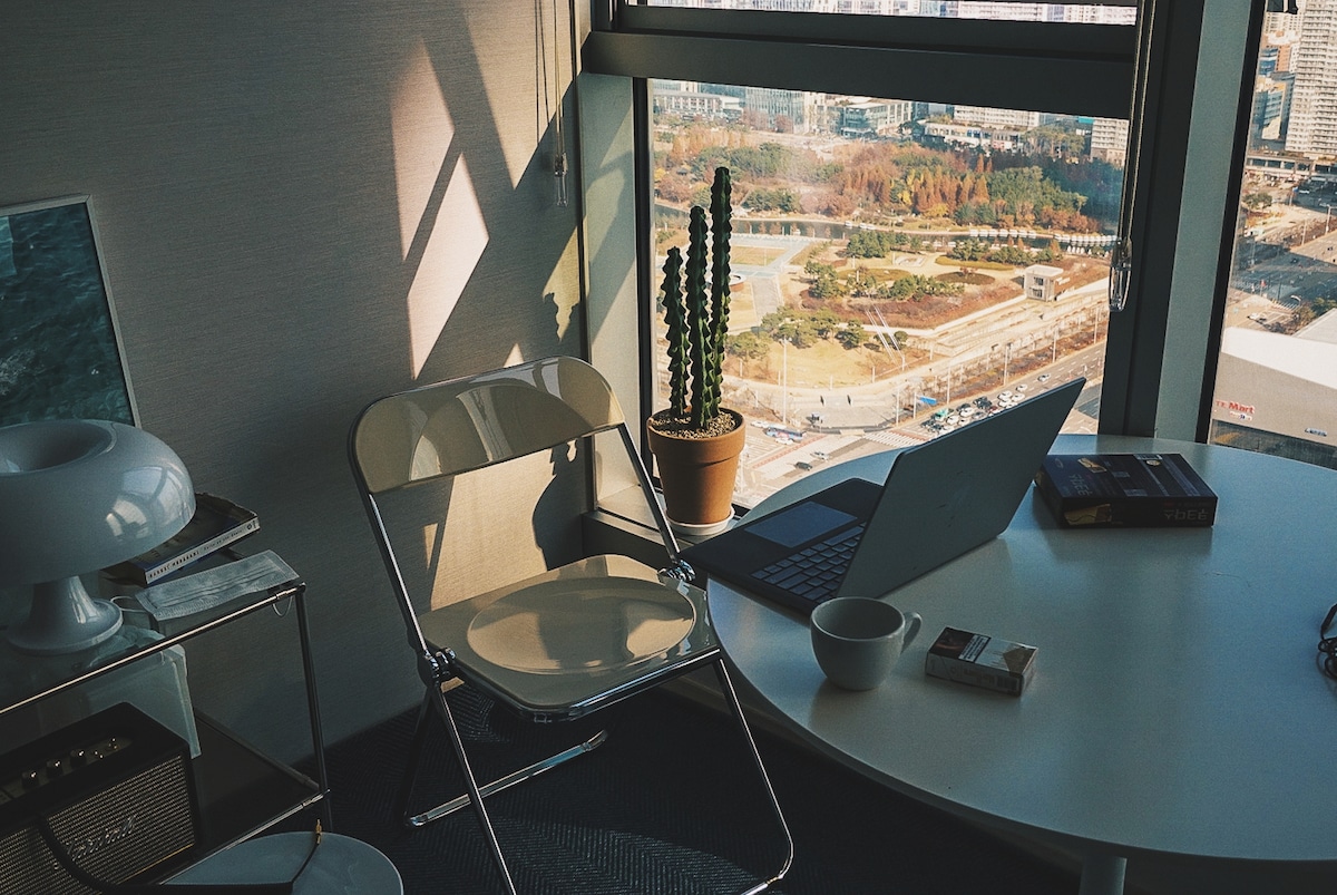 A workspace is set by a large window, featuring a laptop on a round table beside a chair. A potted cactus adds a touch of greenery, while natural light illuminates the area, providing a view of the surrounding landscape.