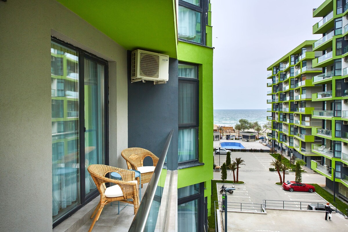 A balcony features two woven chairs positioned next to a glass railing, offering a view of the sea. An air conditioning unit is mounted on the wall, and nearby buildings create a modern architectural backdrop. The tranquil parking area is visible below.