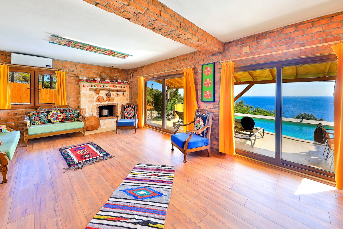 A spacious living area is characterized by exposed brick walls and large windows offering a view of the pool and sea. Colorful textiles adorn the seating, and a cozy fireplace adds warmth to the space. Natural light floods in, creating a welcoming environment.