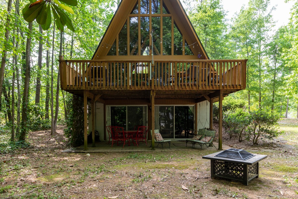 The A-frame cabin is set among trees, featuring large windows that enhance natural lighting. A spacious deck with a railing is visible above, while the ground level includes outdoor seating and a fire pit. The surrounding area is tranquil and landscaped with greenery.