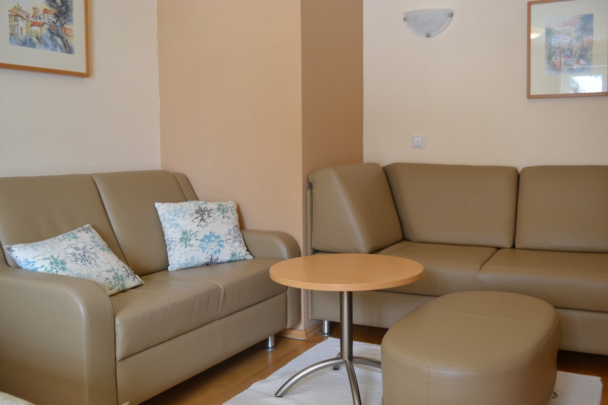 A modern seating area is furnished with a light-colored sectional sofa and a pair of comfortable armchairs. A round wooden table is positioned at the center. Decorative cushions with subtle patterns add a touch of color to the sofa, complementing the warm tones of the walls.