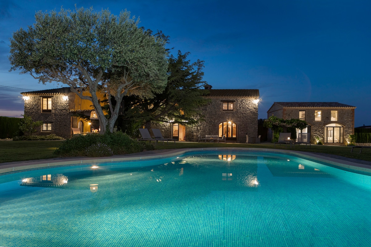 A tranquil outdoor setting is depicted, featuring a clear pool reflecting the evening sky. The stone buildings, illuminated from within, provide a warm glow against the darkening landscape. An olive tree stands nearby, enhancing the serene ambiance of the garden.