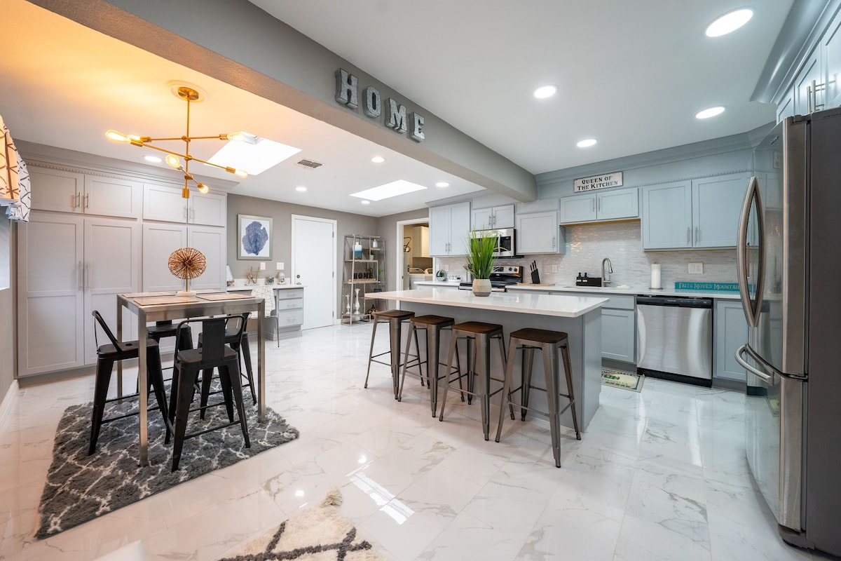 A modern kitchen is presented, featuring a spacious island with bar seating and stainless steel appliances. Gray cabinetry and white quartz countertops provide a sleek look. A dining area with a table and chairs is highlighted, complemented by natural light from overhead skylights.