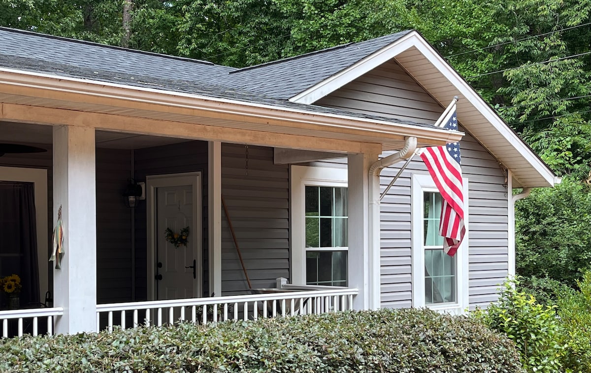 The exterior of the home features a welcoming porch adorned with an American flag. Green hedges frame the entrance, and large windows allow for natural light. The overall design displays a blend of traditional and modern elements, set against a backdrop of trees.
