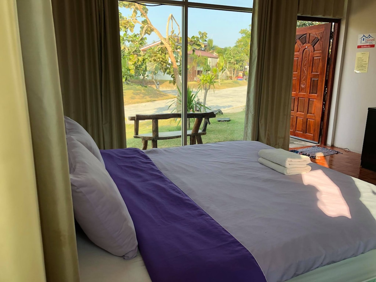 A cozy sleeping area features a spacious bed with purple and white linens. Light streams in through large windows, offering a view of the outdoor landscape. A wooden bench can be seen outside, along with a door leading to the entryway.