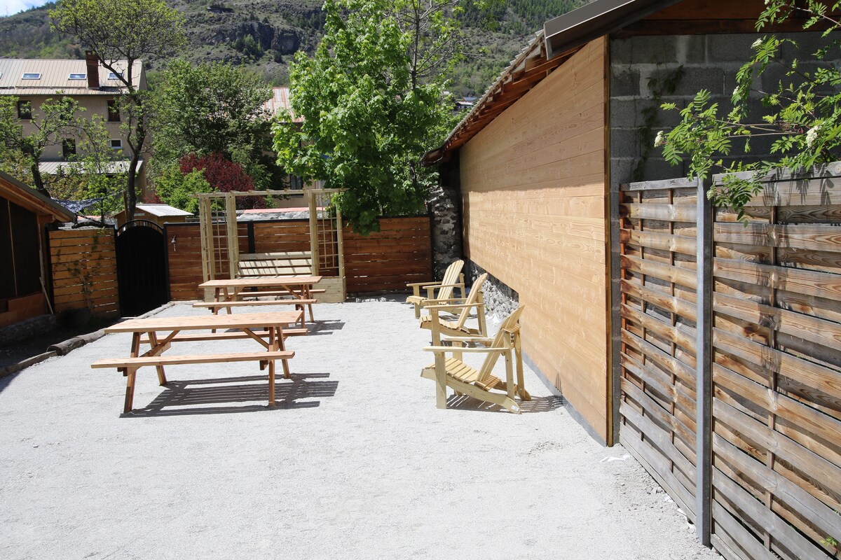 A spacious outdoor area is equipped with several wooden picnic tables and lounge chairs. A wooden frame for swings is positioned against a backdrop of greenery and a rustic wooden wall, creating a relaxed environment for gatherings.