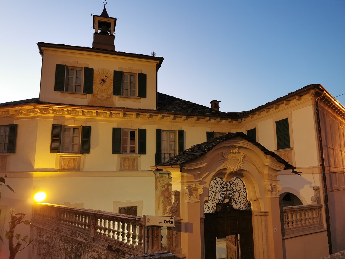 A historic villa is depicted at dusk, featuring elegant architectural details such as decorative moldings and a prominent clock above the entrance. Windows are framed by green shutters, and warm lights illuminate the façade, highlighting the property's grandeur and inviting presence.