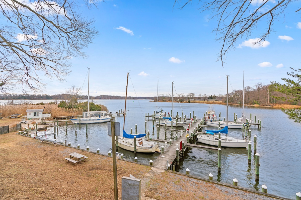 The serene marina setting features several boats moored along wooden docks. Lush greenery lines the shoreline, and calm waters reflect the clear sky. A picnic table is positioned on the grass, offering a perfect spot to relax and enjoy the view.