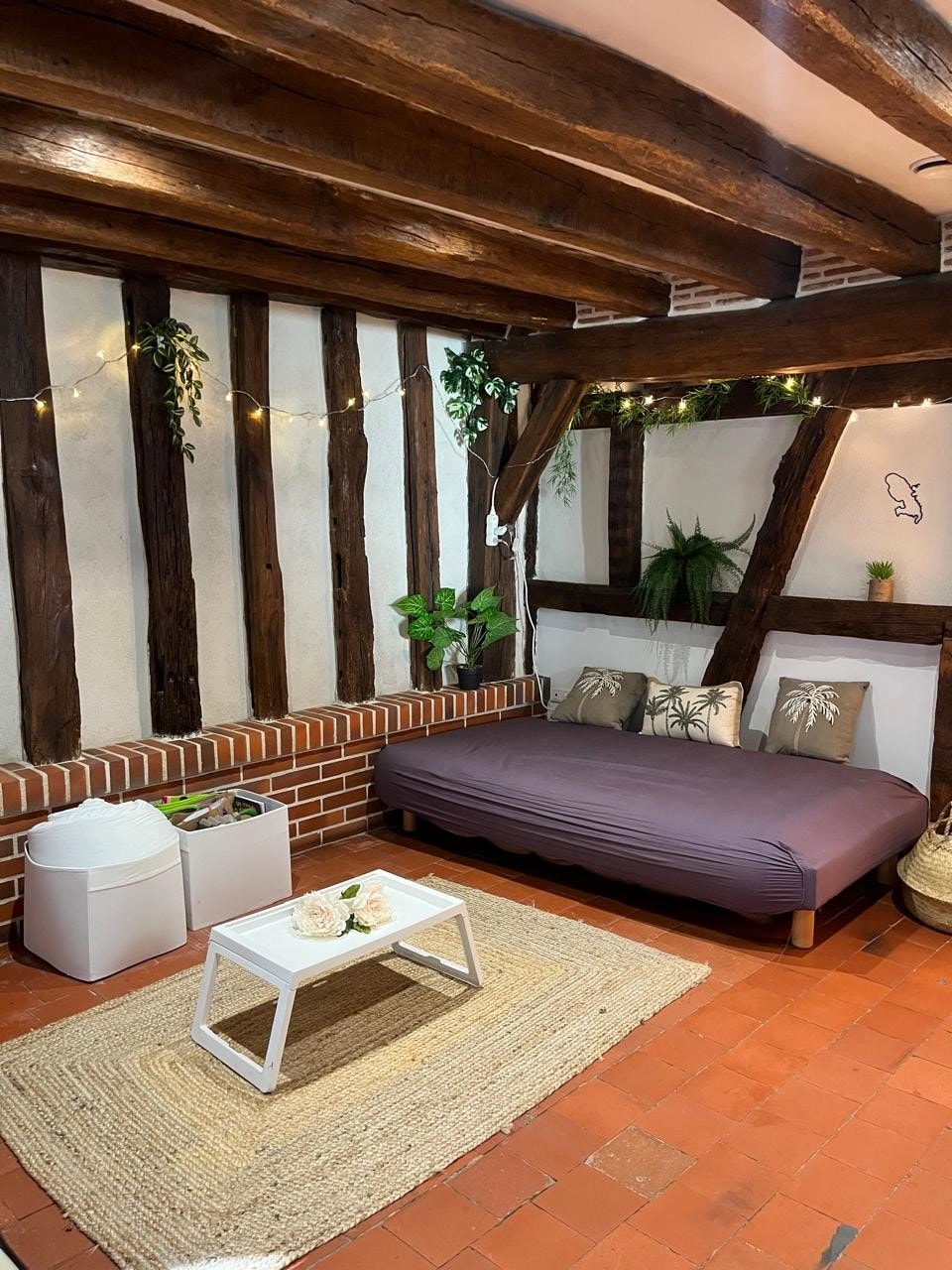A cozy sleeping area features a spacious bed with a purple coverlet, accompanied by decorative pillows. Natural elements, such as plants, are placed along the exposed wood beams. A small white table and a round rug add functionality to the room, creating a relaxed and inviting space.