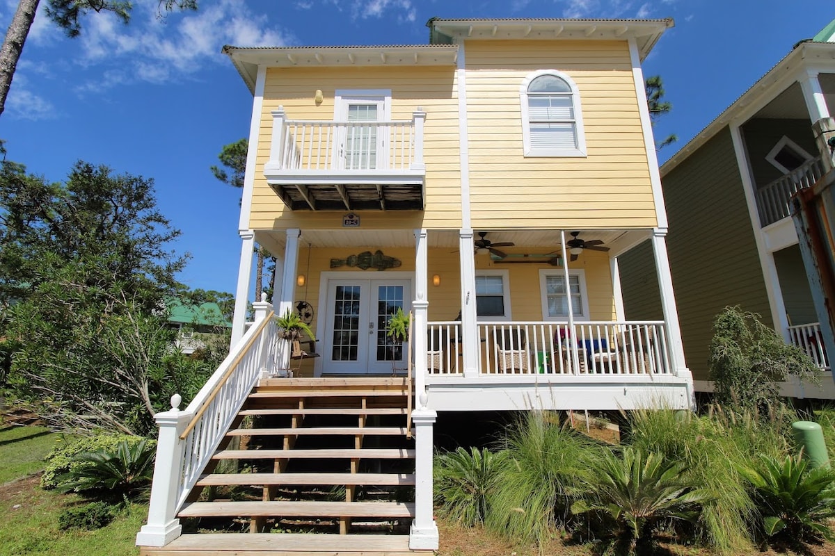 A two-story yellow house is showcased, featuring a wide set of wooden steps leading to the entrance. Large glass doors create a welcoming entry, while a small balcony is seen on the second floor. Lush greenery surrounds the property, enhancing the outdoor appeal.