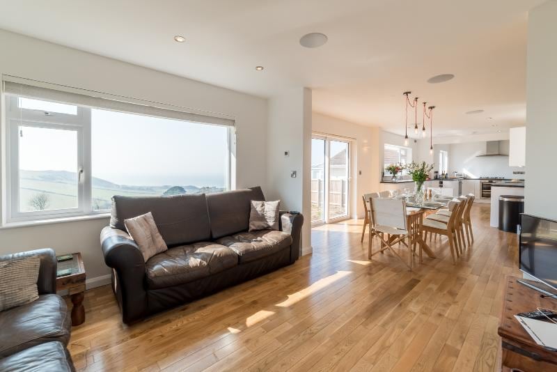 An open plan living space is showcased with a comfortable leather sofa and a rustic coffee table. Large windows provide scenic views, allowing natural light to fill the area. A dining table with wooden chairs is seen in the background, complementing the elegant kitchen layout.