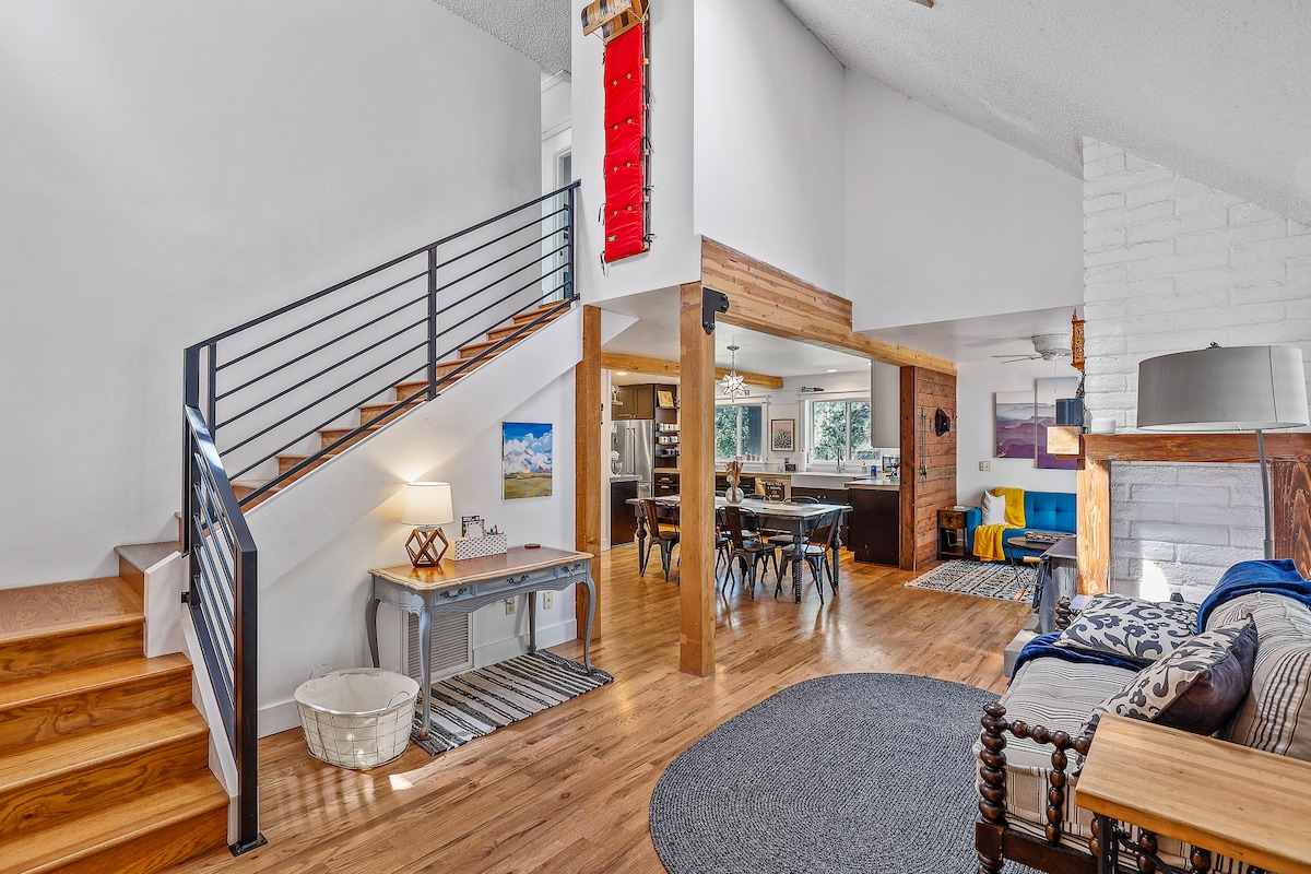 The open living area features a staircase leading to the upper level, with wooden floors and a cozy seating arrangement. A stylish table sits nearby, and the kitchen area is visible in the background, well-lit by large windows that invite natural light.