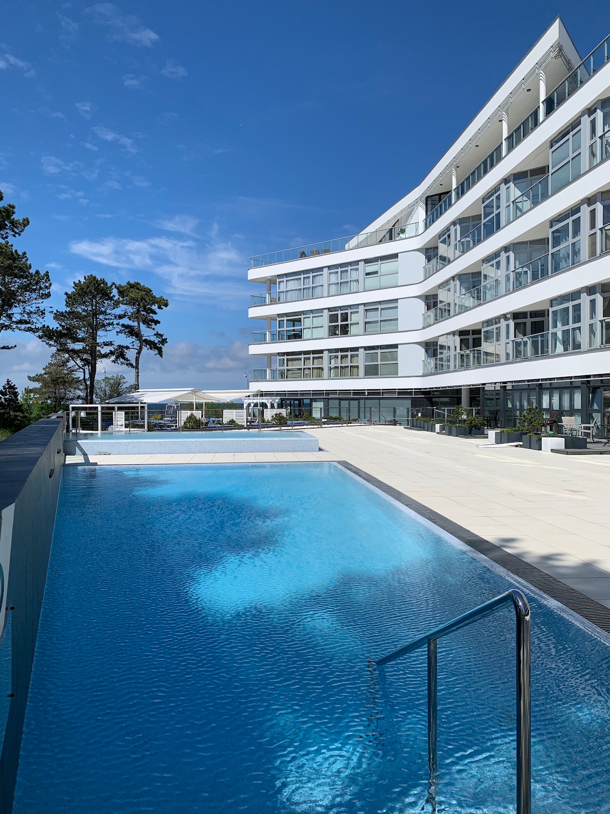 An outdoor pool reflects the clear blue sky, surrounded by a spacious sun terrace. The modern building, featuring large glass windows, is visible in the background. Lush trees add greenery to the scene, enhancing the tranquil setting.