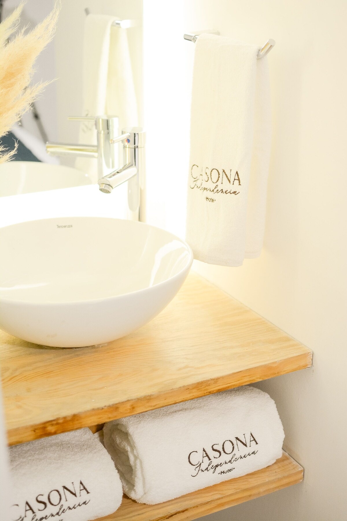 A modern bathroom sink area features a round white basin atop a wooden countertop. Fluffy white towels with 'CASONA' embroidered are neatly arranged on a shelf below. Soft lighting enhances the clean and minimalist design.