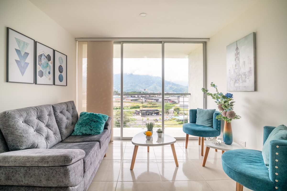 A modern living area is highlighted by large glass doors offering a panoramic view of the surrounding mountains. Plush gray seating is complemented by turquoise accent chairs. A small round table displays decorative items, enhancing the welcoming atmosphere of the space.
