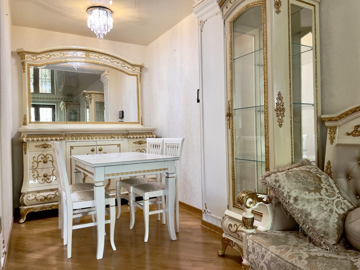 A dining area is featured, showcasing an elegant white dining table accompanied by four chairs. A mirrored cabinet stands against the wall, enhancing the space's light. Decorative details in gold highlight the furniture. A comfortable sofa is positioned nearby, contributing to the overall aesthetic.