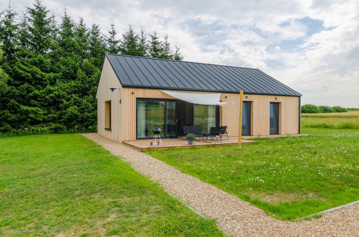 A modern wooden house is set against a backdrop of lush green trees. Large glass doors lead to a wooden terrace, where outdoor seating is arranged. A pathway of gravel extends from the house, surrounded by a grassy area, creating a serene outdoor environment.