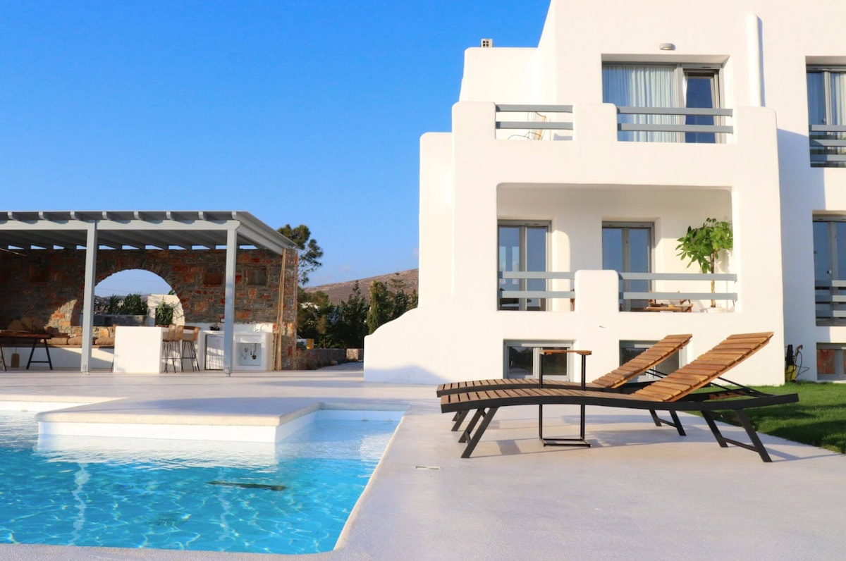 A spacious outdoor area is highlighted by a private swimming pool and sun loungers. A covered outdoor kitchen and dining area are visible, providing a shaded space for gatherings. The villa's clean lines and modern architecture contrast with the clear blue sky.