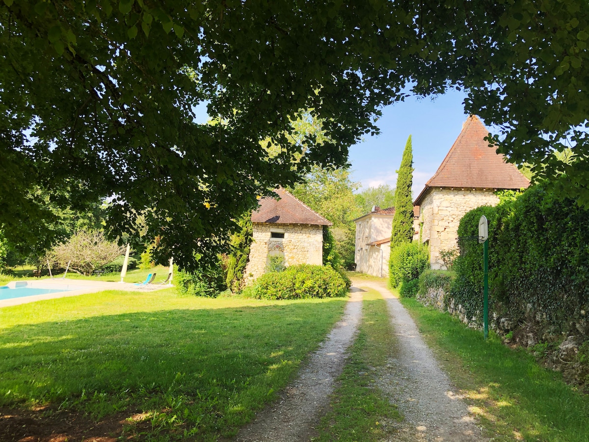 A gravel path lined with lush greenery leads to a charming stone villa complex. The inviting pool is visible on the left, surrounded by a well-maintained lawn. The buildings feature traditional roofs and a serene setting, offering a glimpse of the tranquil landscape.