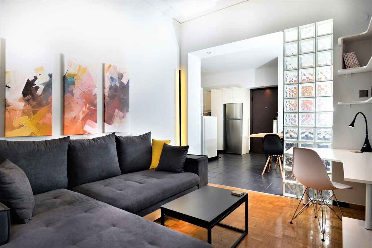 The living area displays a modern sofa in soft gray, complemented by abstract art pieces on the wall. A sleek black coffee table stands in front of the sofa. A well-lit kitchen is visible in the background, featuring contemporary appliances and a dining area with simple furniture.