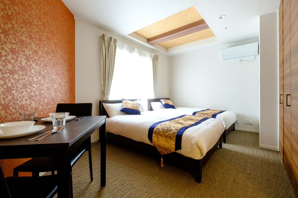 A spacious room features two double beds, adorned with elegant fabrics and accented by textured wallpaper in a warm hue. Natural light streams through a large window, while a small dining table and chairs are positioned nearby, enhancing the functional design.