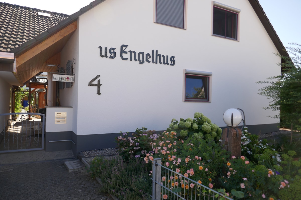 The entrance of the property is presented, featuring a white exterior wall with black lettering displaying the name 'us Engelhus.' A garden area is visible, adorned with vibrant flowers and greenery, creating a welcoming approach to the residence.