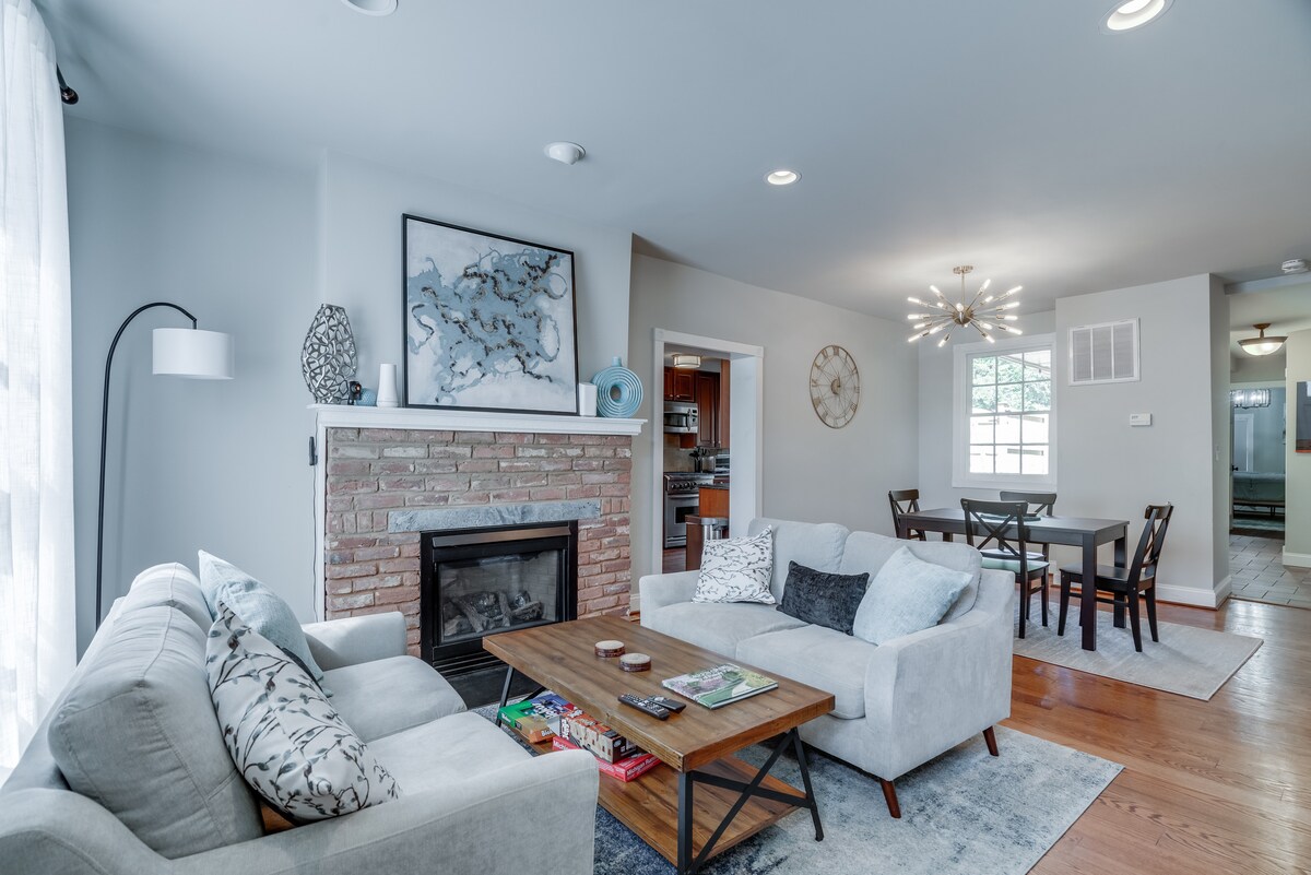 A comfortable living area features two light grey sofas arranged around a wooden coffee table. A brick fireplace serves as a focal point, complemented by a wall-mounted artwork. A dining area is visible in the background, along with a window allowing natural light.