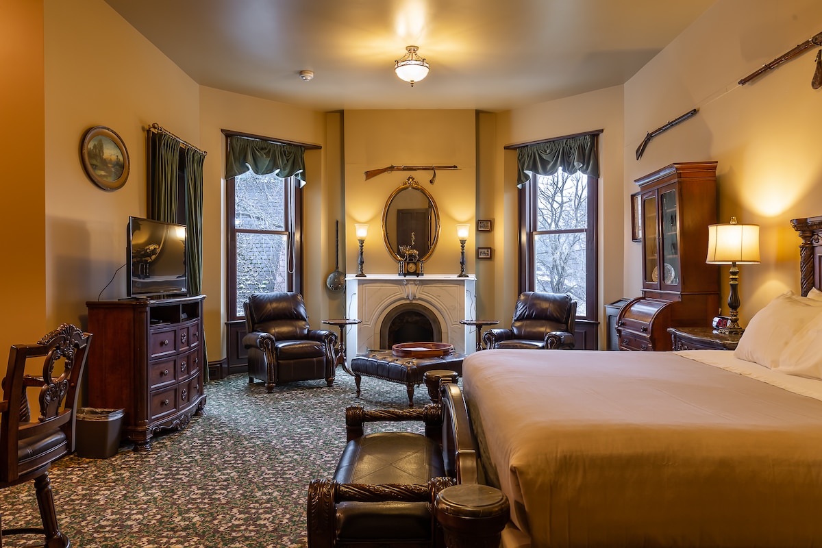 A spacious room is furnished with a large bed, polished wooden furniture, and two armchairs positioned near a fireplace. Large windows allow natural light to fill the space, complementing the warm wall colors and elegant decor. A television is mounted on a dresser for entertainment options.