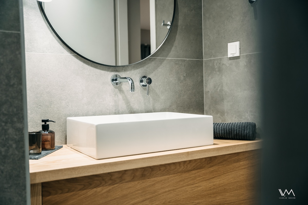 A modern bathroom features a rectangular vessel sink on a wooden countertop, complemented by a round mirror above. Minimalistic faucets are mounted on the wall, and personal care items are placed nearby for convenience.