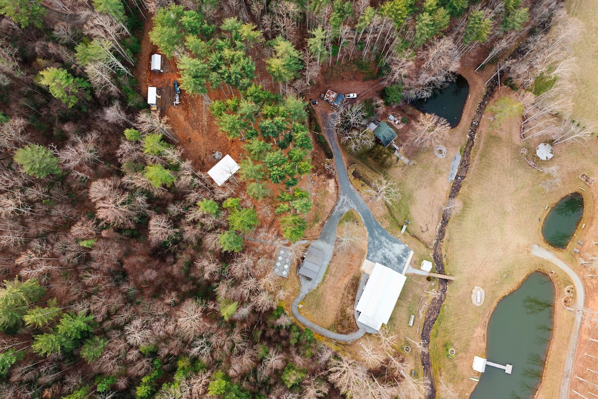 An aerial view captures the layout of the property, showcasing a mix of wooded areas and open spaces. Visible are the farmhouse, a gravel path, two ponds, and several outdoor structures nestled among the trees, highlighting the natural surroundings.