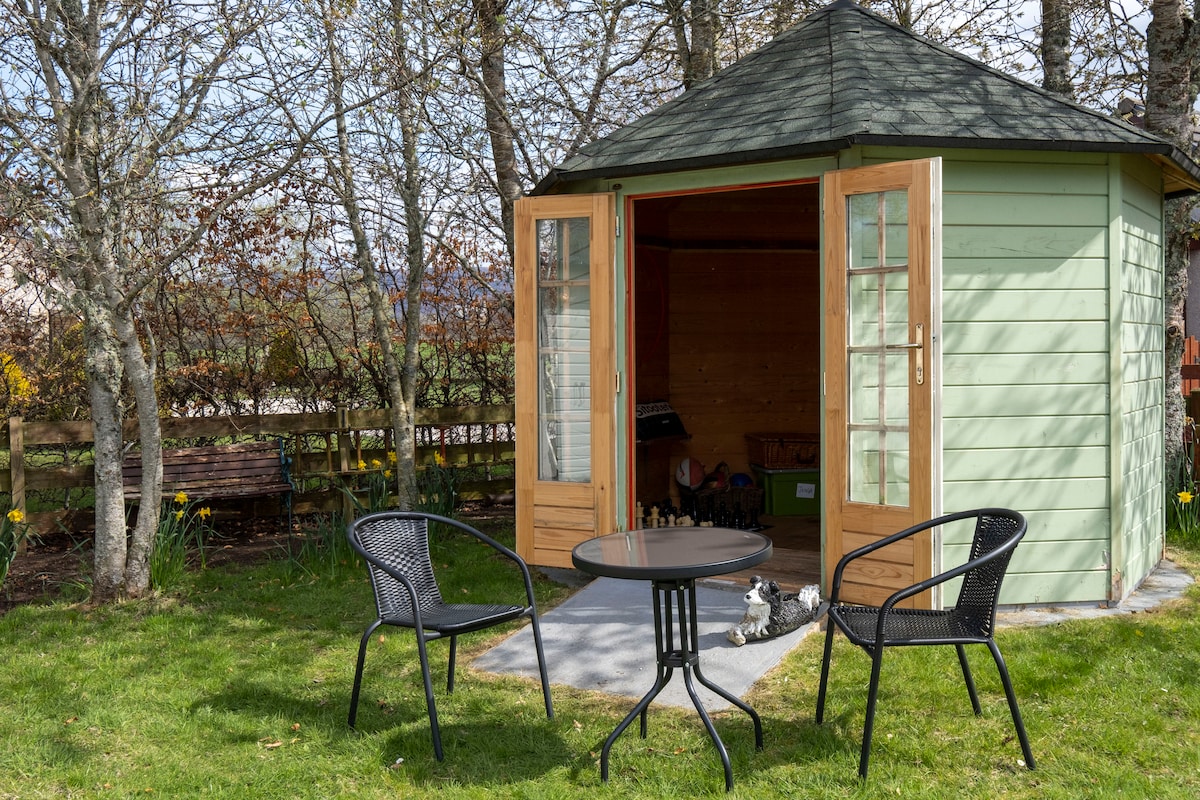A garden structure featuring large glass doors is surrounded by greenery and trees. Two black chairs are positioned at a small table outside, creating a space for relaxation. The interior appears to be a cozy area, with wooden paneling visible through the open doors.