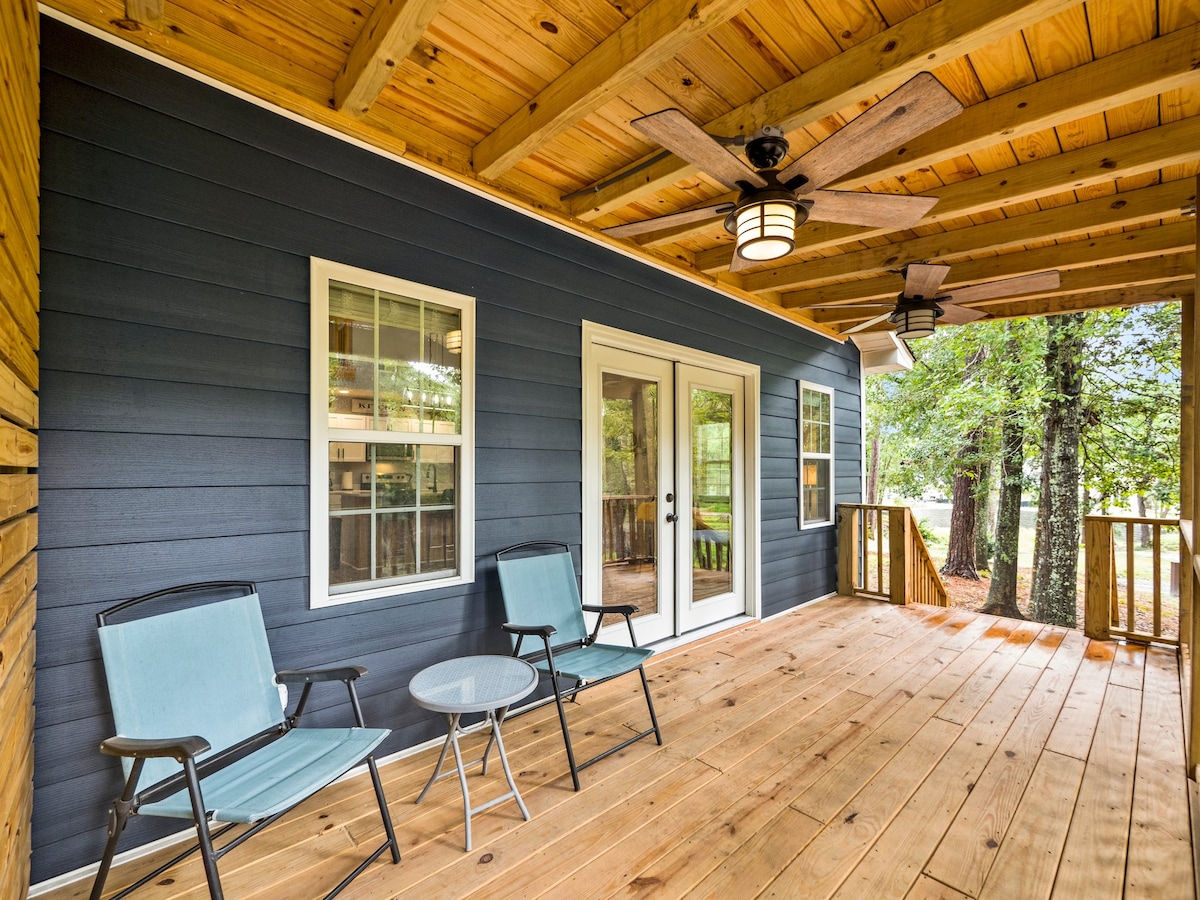 A covered porch area features two light blue chairs and a small round table on weathered wooden flooring. Sliding glass doors lead indoors, framed by white trim, while overhead fans provide ventilation. The porch is surrounded by trees, enhancing a sense of openness.