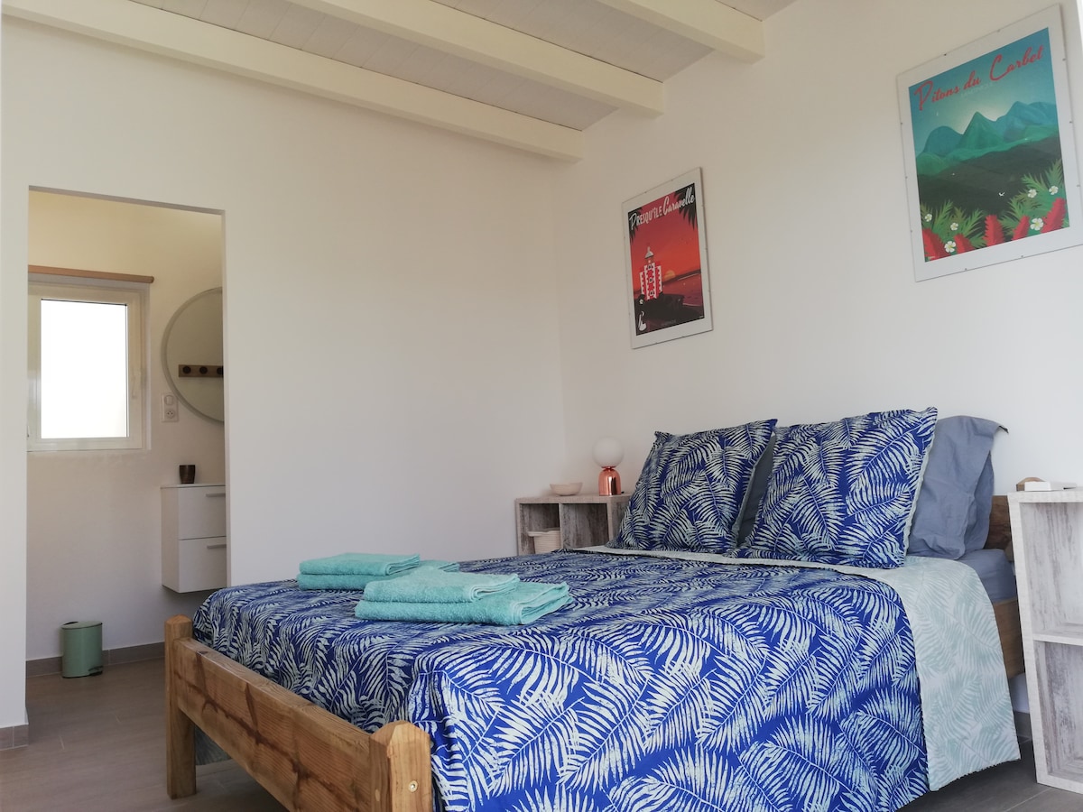 A spacious sleeping area is presented with a wooden bed featuring a leaf-patterned blue duvet cover. Soft, turquoise towels are neatly arranged at the foot of the bed. Light enters through a nearby window, complementing the minimalist decor and artwork on the walls.