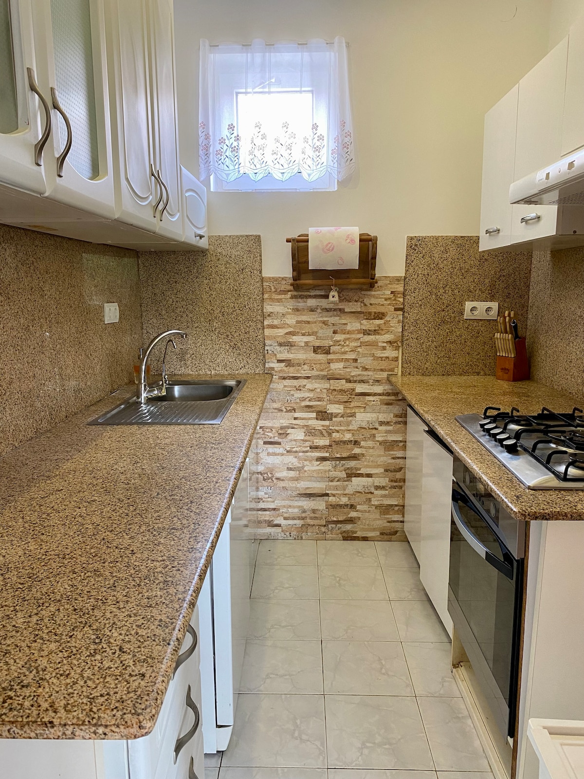 A compact kitchen features granite countertops and a backsplash of light-colored stones. Cabinets with glass fronts provide storage, while a sink and a gas stove are positioned thoughtfully along the wall. Natural light enters through a window adorned with lace curtains.