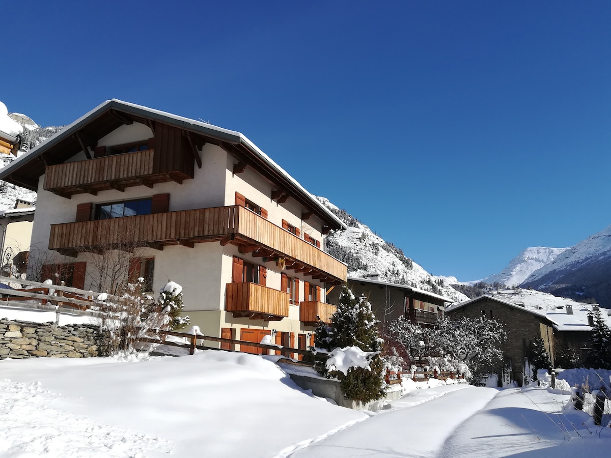 A charming village house is set against a backdrop of snow-covered mountains. The building features wooden balconies and warm tones, surrounded by a blanket of fresh snow. The clear blue sky above enhances the serene winter atmosphere.
